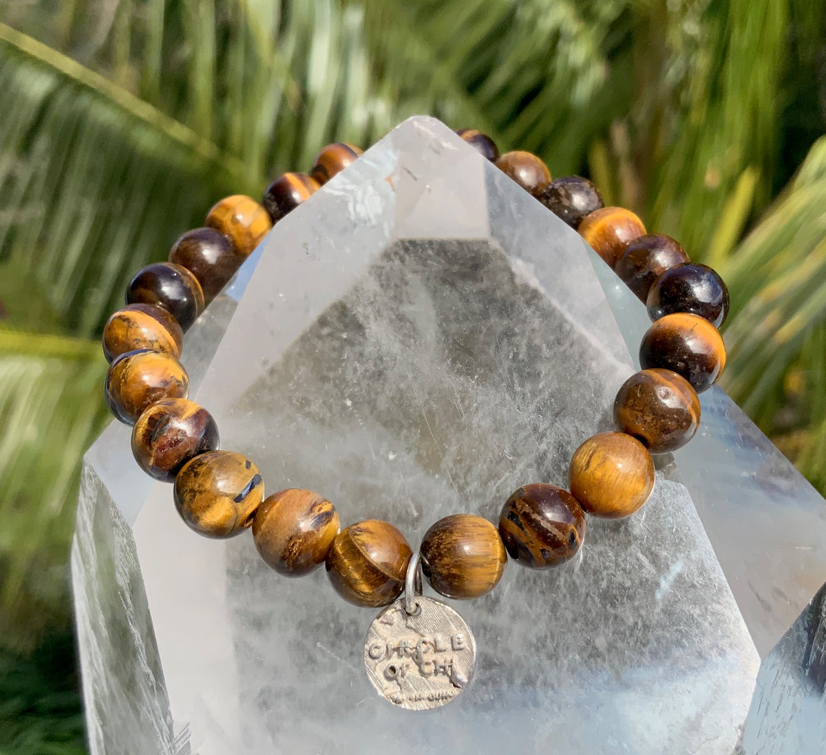 A bracelet with a series of brown and beige beads, featuring a tiger's eye effect, displayed on a crystal stand in a tropical setting.